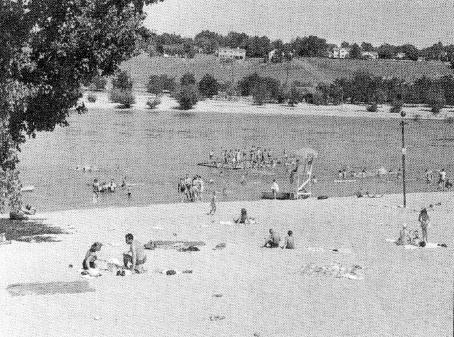 Black and white photo of a sandy beach with visitors swimming and on a raft, trees and houses visible on distant hillside