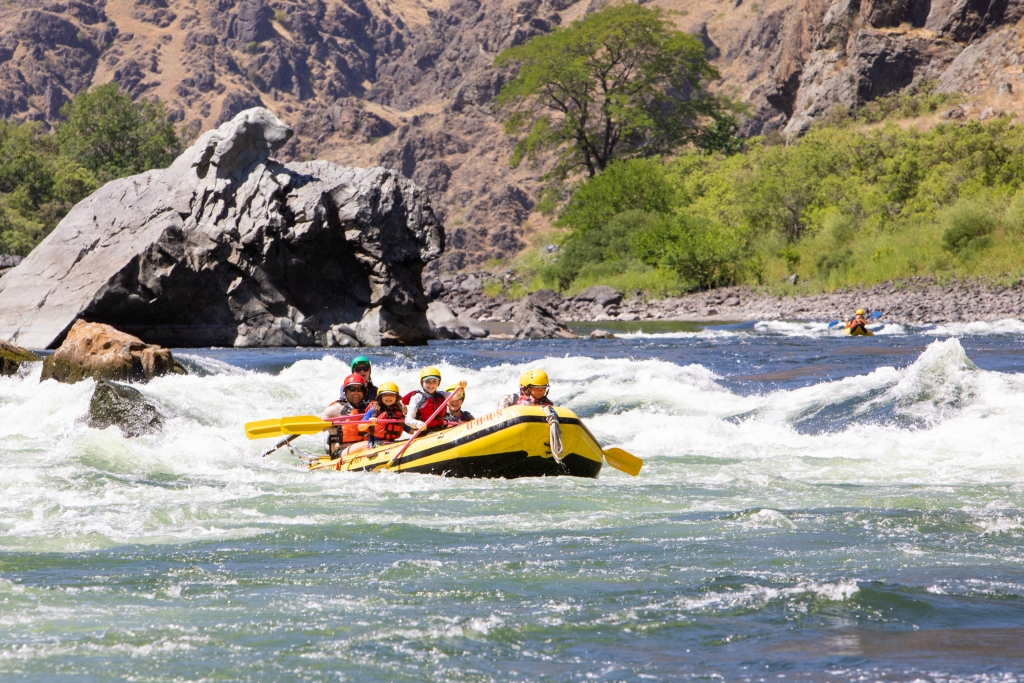 Four people in a yellow whitewater raft navigate rapids on a rocky river surrounded by canyon walls and green vegetation