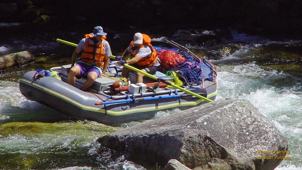 Three people in orange life jackets navigate a whitewater raft through turbulent rapids near large rocks