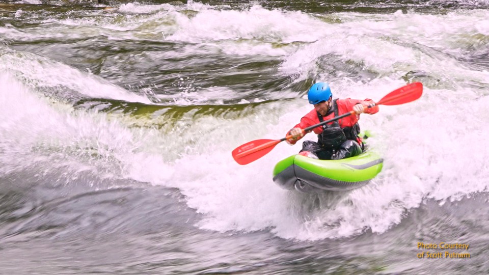 Kayaker in red shirt and blue helmet paddles bright green kayak through white water rapids