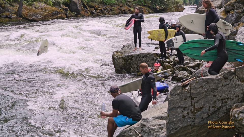 River surfers in wetsuits prepare boards by rocky shore with rushing white water and forested banks