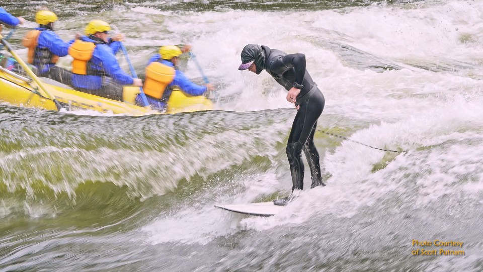Surfer in black wetsuit riding wave with yellow raft and blue-shirted paddlers in background