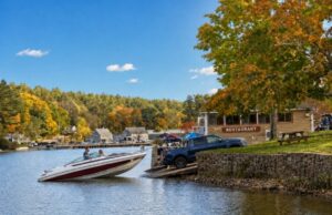 Speedboat at community boat ramp with restaurant building, blue truck, and autumn foliage along waterfront