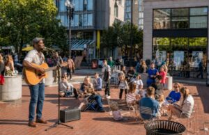 Man playing acoustic guitar at outdoor community concert in brick plaza with seated audience and modern buildings